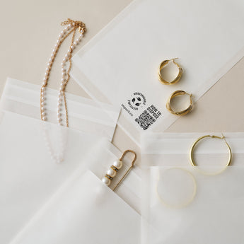 Several impack.co Glassine Bags 5.6" x 8.6" containing golden hoop earrings and pearl necklaces are laid out neatly on a beige surface.