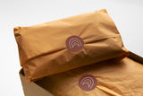 Two brown paper packages sealed with Custom Paper Sticker Sheets from impack.co, featuring an abstract white design on uncoated paper. One package is placed inside a cardboard box, while the other rests on top.