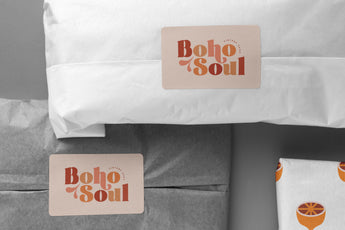 Packages and wrapped items labeled with "Boho Soul" stickers are arranged in a flat lay. The personalized packaging includes white and gray materials, along with one item wrapped in fruit-patterned paper. The Custom Paper Sticker Sheets - Single design from impack.co add a unique touch to the presentation.