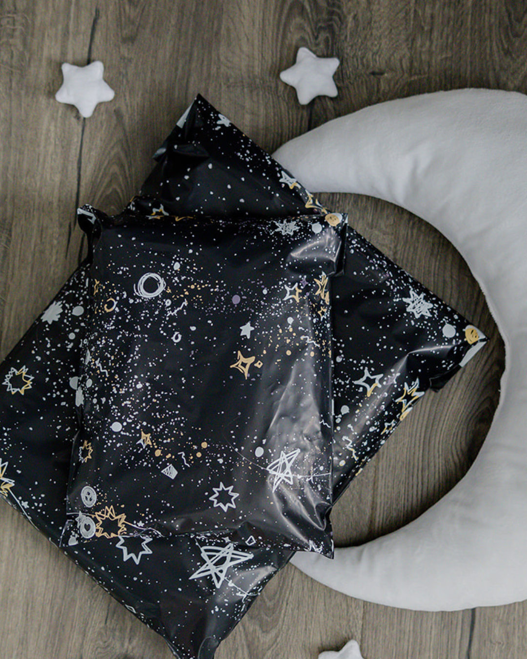 Two black packages with white and gold star and moon patterns rest on a wooden floor next to a plush crescent moon and two small white star pillows.