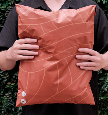 A person in a dark shirt holds a large, orange, patterned package with both hands in front of green foliage.