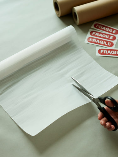 A hand cuts a sheet of packing paper with scissors, next to rolls of brown paper, twine, and red "FRAGILE" stickers on a table.