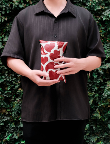 A person in a black shirt holds a gift bag decorated with red heart prints, standing in front of a leafy green background.