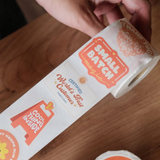 A hand unrolls impack.co's Sticker Roll - Assorted Designs, showcasing colorful, eco-friendly labels like "Small Batch Magic," "World's Best Customer," and "Cool Thing Inside.