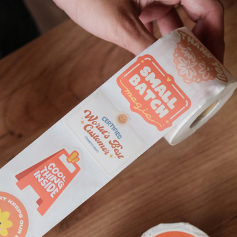A hand unrolls impack.co's Sticker Roll - Assorted Designs, showcasing colorful, eco-friendly labels like "Small Batch Magic," "World's Best Customer," and "Cool Thing Inside.