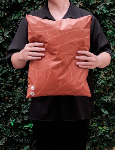 A person in a black shirt holds a large, orange plastic bag with a geometric pattern, standing in front of a leafy green background.