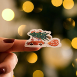 A hand with painted nails holds an impack.co Christmas Thank You Sticker featuring a car with a tree and “HO HO HO”—perfect for holiday packaging, with blurred festive lights in the background.