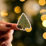 A hand with dark red nail polish holds an impack.co Christmas Thank You Sticker featuring a decorated tree, with blurred golden lights in the background—ideal for festive gift wrap or holiday packaging.