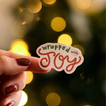 A hand with painted nails holds a Christmas Thank You Sticker from impack.co, featuring the message "wrapped with joy," against a softly blurred background of yellow lights.