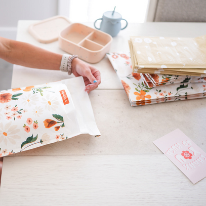 Person assembling floral-patterned padded envelopes on a table, with a partially visible card and a baby blue sippy cup nearby.