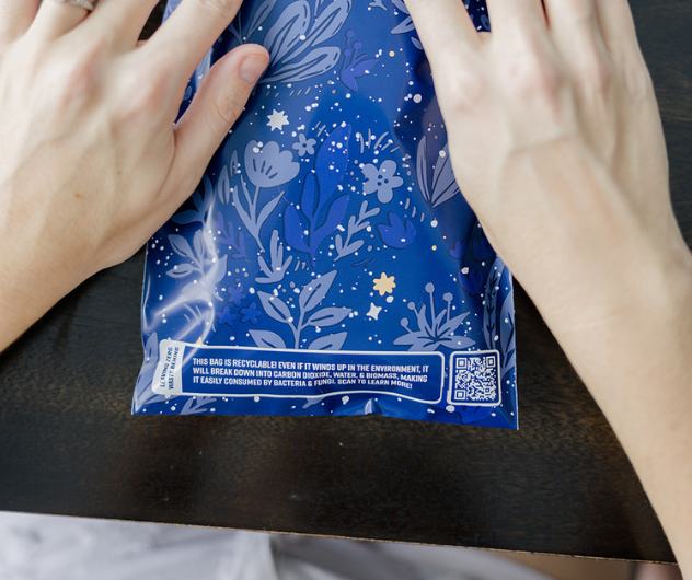 Hands opening a blue patterned package with a recycling message and QR code on a dark surface.