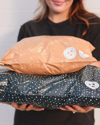 A person holds two patterned plastic mailer bags, one orange and one navy with white dots, against a neutral background.