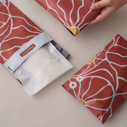 Three packets with red and white abstract designs, one being held and another partially opened to reveal a white inner pouch on a light surface.
