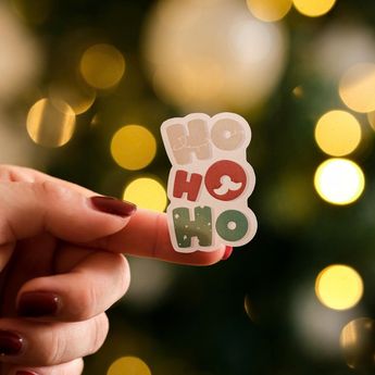 A hand with maroon nail polish holds an impack.co Christmas Thank You Sticker in festive colors—perfect for holiday packaging—with blurred holiday lights in the background.