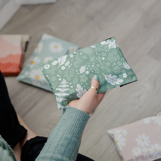 A person holding a green pouch with a white floral pattern, sitting on a floor with other patterned pouches scattered around.