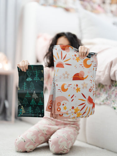A child in pajamas kneels on a carpet, holding impack.co’s 10"x13" Boho Sun & Moon Biodegradable Mailers—an eco-friendly, eye-catching alternative to standard mailers, ideal for stylish boho packaging.