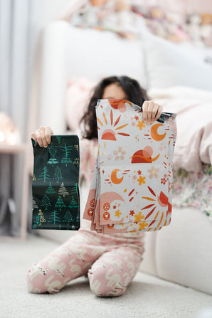 A child in pajamas kneels on a carpet, holding impack.co’s 10"x13" Boho Sun & Moon Biodegradable Mailers—an eco-friendly, eye-catching alternative to standard mailers, ideal for stylish boho packaging.