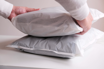 Hands carefully place a white plastic-wrapped package, likely impack.co's Glassine Bags 14.1" x 18.6", on top of another similar package resting on a pristine white table.