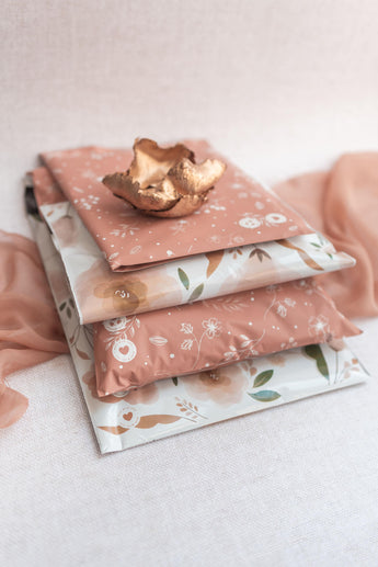 Three impack.co SALE Floral 2D Rosy Brown Biodegradable Mailers (10" x 13") are stacked on a light surface, showcasing eco-friendly, stylish packaging and reliable protection, topped with a decorative gold ceramic dish.