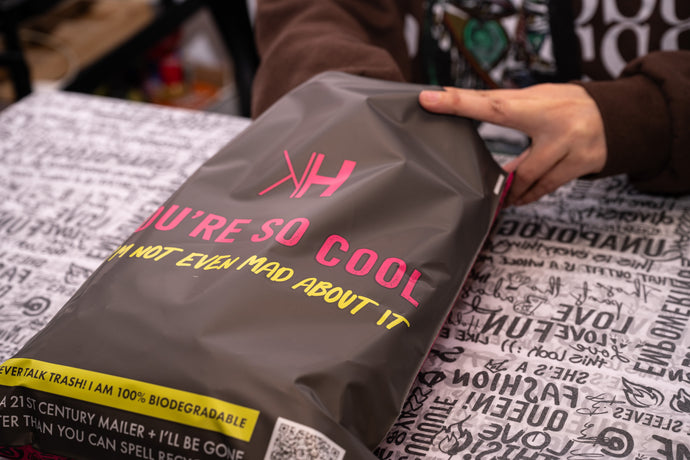 A person wraps an item in a biodegradable mailer with the text "You're so cool, I'm not even mad about it" on a surface covered with various printed patterns.