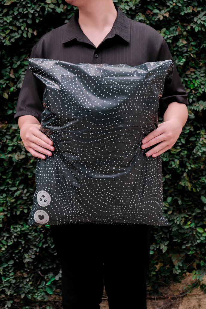 A person in a black shirt holds a large black pillow with a white dotted pattern in front of a green leafy background.