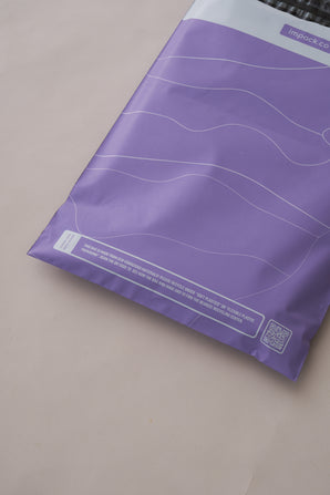 The beige surface features impack.co's SALE - Tidal Lilac Mailers, measuring 10" x 13", adorned with white wavy lines and a QR code. These purple recyclable bags offer versatile packaging solutions.