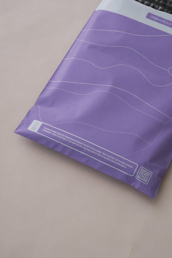 The beige surface features impack.co's SALE - Tidal Lilac Mailers, measuring 10" x 13", adorned with white wavy lines and a QR code. These purple recyclable bags offer versatile packaging solutions.