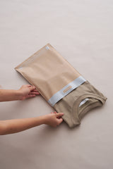 Hands are seen sliding a folded beige T-shirt into a Tidal Sand Mailers 10" x 13" by impack.co with a translucent window, on a light-colored surface. This versatile packaging solution ensures both visibility and transit protection.