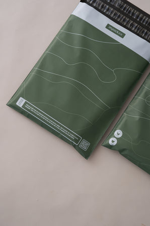Two green, eco-friendly mailing bags with white line designs, one partially shown. The Tidal Moss Mailers 10" x 13" from impack.co, ideal for medium-sized shipments, feature branding and a QR code. Made from biodegradable materials, they offer reliable transit protection on a beige surface background.
