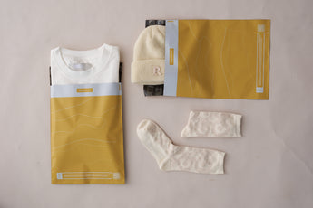 A folded white t-shirt, a beige knit hat, and patterned socks rest beside impack.co's Tidal Amber Mailers 10" x 13", nestled on a white background.