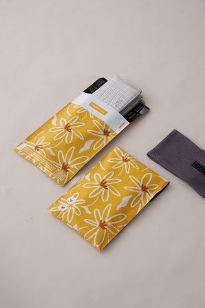 Two yellow SALE - Golden Pencil Floral Mailers 6" x 9" by impack.co, one partially opened with two gray items inside, and a folded gray fabric item nearby. These biodegradable mailers add charm to everyday transit protection for those seeking sustainable options.