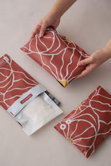 Hands holding an impack.co sale item: Crimson Grove Mailers 6" x 9", red with white abstract lines, beside two more on a surface. One pouch is open, showing its contents.