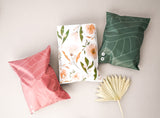 The pink, floral, and green decorative pillows with leaf designs rest on a light surface beside a graceful dried leaf. They arrived intact in versatile "Olive Leaf Biodegradable Mailers" by impack.co, ready to charm any space.