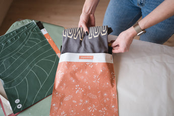 Someone gently places a folded shirt into an Olive Leaf Biodegradable Mailer by impack.co on the table, with more mailers ready to go.