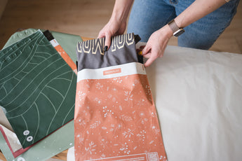 A person places a folded t-shirt into an impack.co SALE - Floral 2D Rosy Brown Biodegradable Mailer (10" x 13") on a table, with other eco-friendly mailers nearby, highlighting versatile and protective shipping solutions.