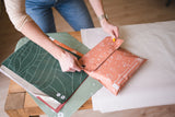 A person seals an impack.co SALE - Floral 2D Rosy Brown Biodegradable Mailer (10" x 13") at a table with green mailers and white tissue paper, showcasing versatile eco-friendly packaging options.