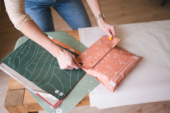 A person seals an impack.co SALE - Floral 2D Rosy Brown Biodegradable Mailer (10" x 13") at a table with green mailers and white tissue paper, showcasing versatile eco-friendly packaging options.