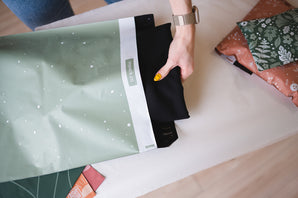 Someone puts a black garment into a SALE - Starry Lily Pad Biodegradable Mailer by impack.co on a table surrounded by folded clothes, ensuring protection during transit.