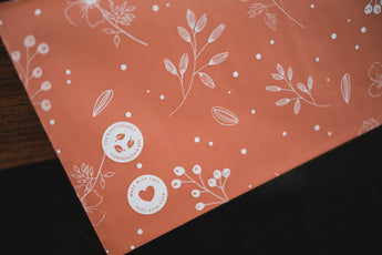 A close-up of impack.co's SALE - Floral 2D Rosy Brown Biodegradable Mailers (10" x 13") shows white floral patterns and stickers reading "Made with Love," adding an eco-friendly, charming touch to your packaging.