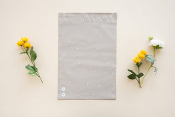A SALE sandstone biodegradable mailer by impack.co, sized 10"x13" in beige with a subtle polka dot pattern, is showcased between two small floral arrangements on a light cream background.