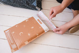 Person sealing an 8.5" x 12" SALE - Celestial Tan Biodegradable Bubble Mailer from impack.co on a white wooden floor.