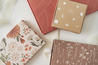 Four decorative gift boxes, including one impack.co ABC Box Cedar Leaf 9.8" x 13.3" - Extra Large, showcase various floral and celestial patterns arranged on a white surface. Cream ribbon is partially visible in the arrangement.