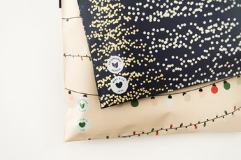 Two overlapping sheets of wrapping paper: one with gold and white dots on black, another with festive lights on cream, ideal for holiday designs. Leaf design stickers are visible. Consider impack.co's SALE - Christmas Tree Biodegradable Mailers 10" x 13" to match your festive needs.