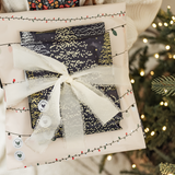 A person holds an impack.co SALE - Christmas Light Strings Biodegradable Mailer (10" x 13" US), wrapped with a white ribbon, while standing by a sparkling Christmas tree.