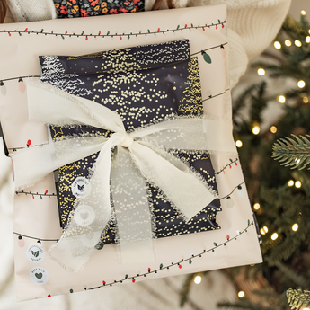 A person holds a SALE - Christmas Light Strings Biodegradable Mailer 14.5" x 19" by impack.co, wrapped festively with a white ribbon, near a lit and decorated Christmas tree.