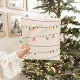 A person holds a SALE - Christmas Light Strings Biodegradable Mailer (10" x 13") by impack.co, featuring festive string light and snowflake designs, in front of a decorated Christmas tree and framed photos.