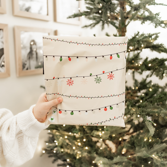 A hand holds an impack.co SALE - Christmas Light Strings Biodegradable Mailer (14.5" x 19" US) with festive string lights and snowflake designs, in front of a lit Christmas tree.