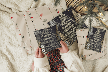 A child holds a wrapped gift with tree patterns, nestled among more presents and a basket on a soft surface. impack.co's SALE - Christmas Tree Biodegradable Mailers 6" x 9" CA ensure safe transit for all festive surprises under the tree.