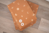 A stack of impack.co's SALE - Celestial Tan Biodegradable Bubble Mailers 8.5" x 12" CA sits on the wooden floor, featuring orange celestial designs and eco-friendly materials. These mailers provide stylish, sustainable cushioning for all your shipping needs.