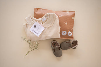 Folded baby clothes with a tag rest beside an eco-friendly brown envelope and white flowers. Gray baby shoes complete the scene on a light background, cushioned by impack.co's SALE - Celestial Tan Biodegradable Bubble Mailers 8.5" x 12" CA for gentle delivery.
